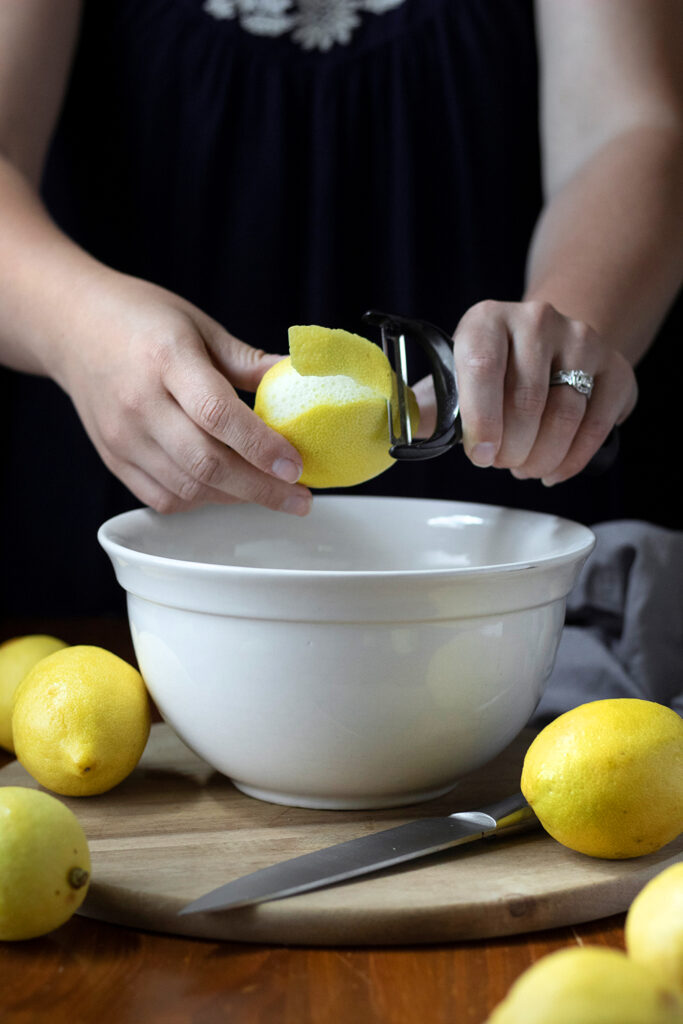 using a vegetable peeler to remove lemon peels over a mixing bowl.