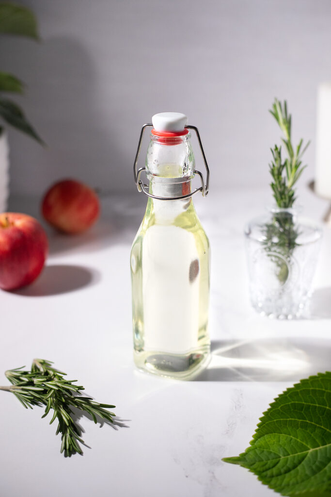 a swing top bottle filled with pale green syrup next to rosemary sprigs and apples.