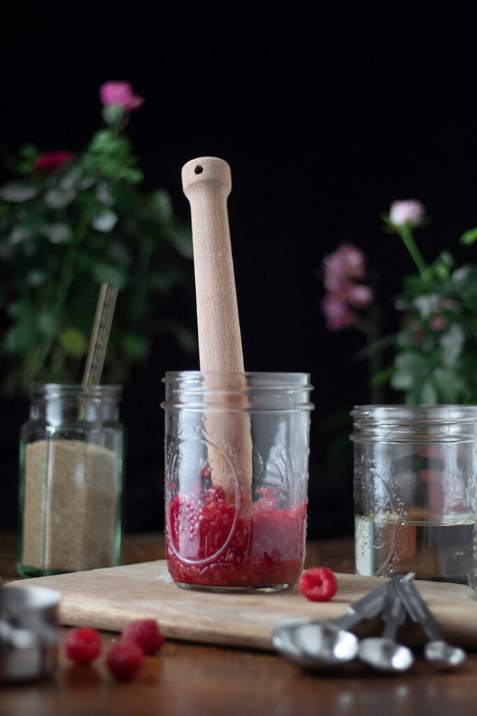 a wooden muddler in a mason jar full of mashed raspberries.