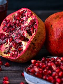 Pomegranate seeds in a bowl next to a whole pomegranate and a pomegranate cut in half